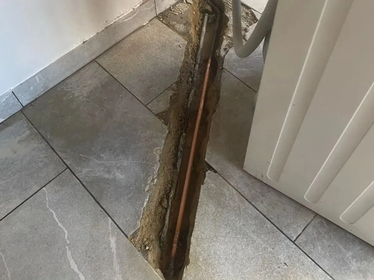 A narrow opening in grey utility-room floor tiles exposes copper pipe beside an appliance