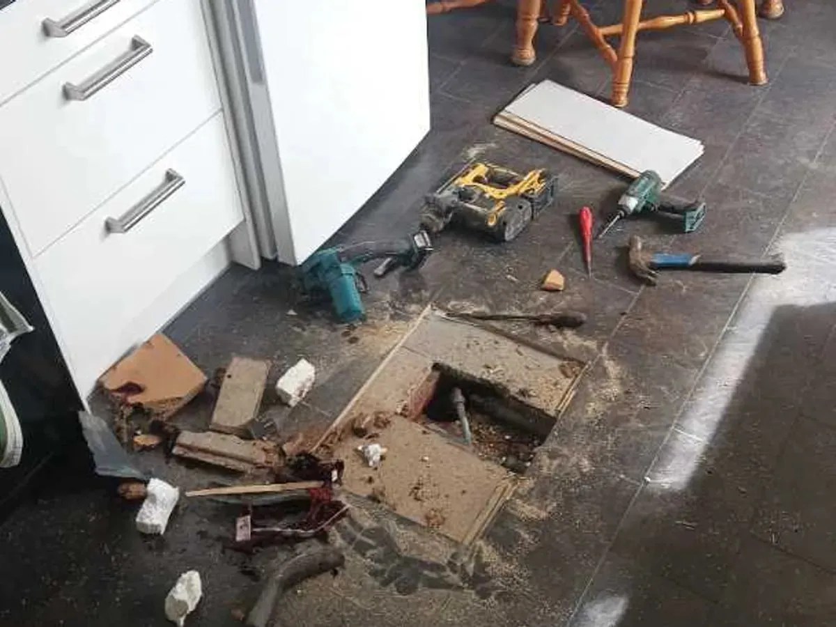 A small rectangular opening in the kitchen floor exposes pipework beside the base units